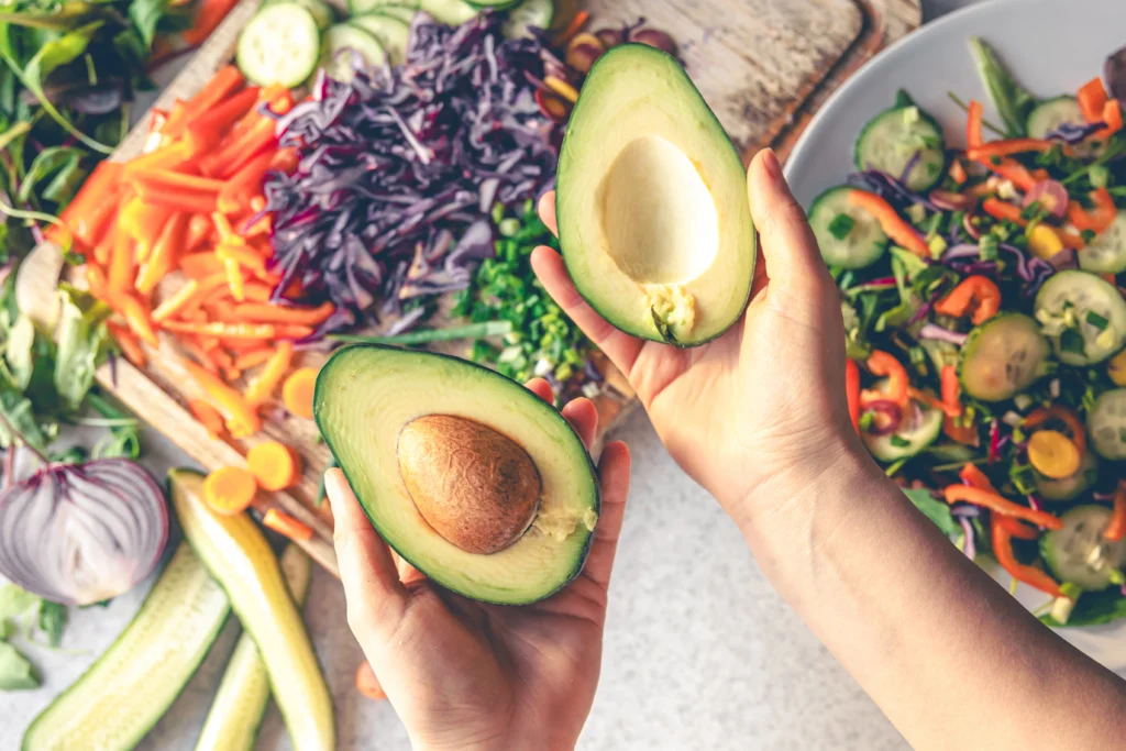 Mãos femininas segurando um abacate, com vegetais ao fundo, representando uma alimentação rica em gorduras saudáveis.