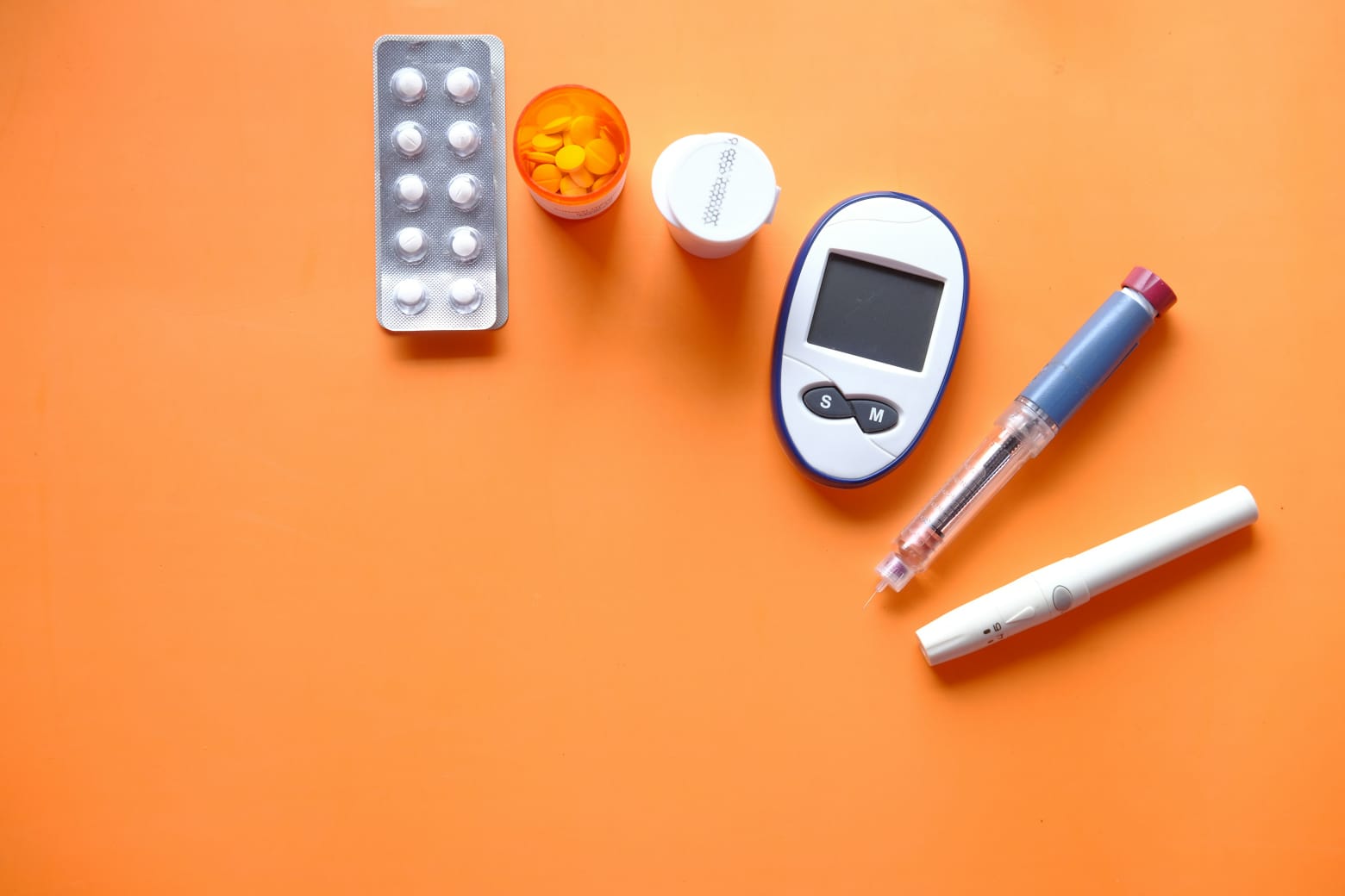 Kit de controle do diabetes com medicamentos, glicosímetro, seringa de insulina e lanceta sobre fundo laranja.