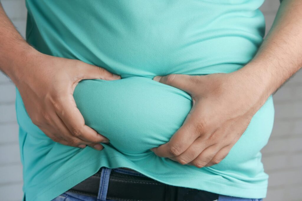 Pessoa apertando a barriga por cima da camiseta, representando o acúmulo de gordura abdominal relacionado à obesidade.