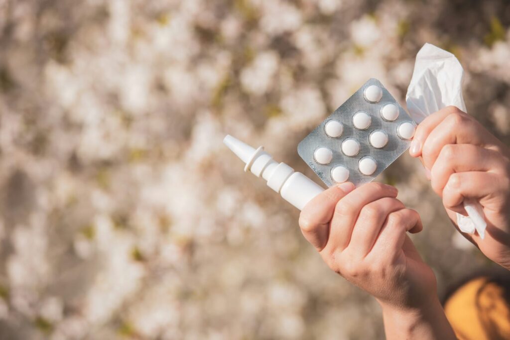 Mãos seguram cartela de comprimidos, spray nasal e lenço, representando o uso de de multiplos medicamentos, chamado de polimedicação.