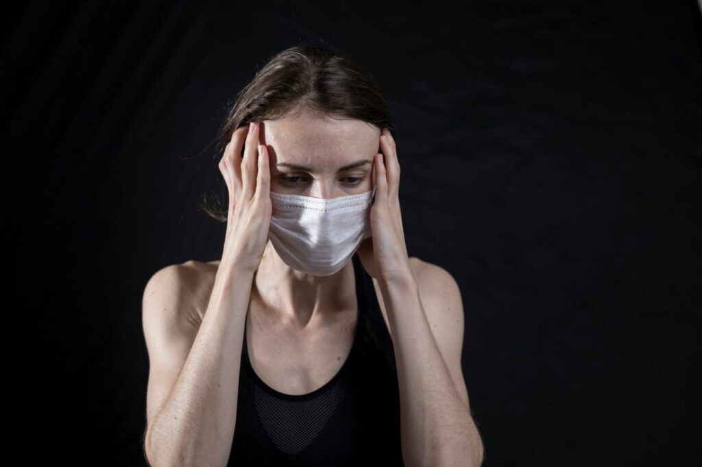 Mulher usando máscara e pressionando a cabeça com as mãos, demonstrando dor facial comum em casos de sinusite.