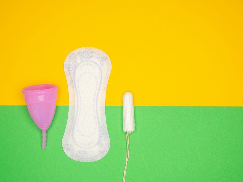 Coletor menstrual, absorvente e tampão lado a lado sobre fundo colorido, representando cuidados no período da cólica menstrual.