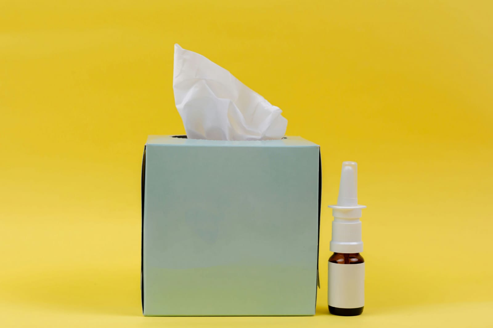 caixa de lenços e frasco de spray nasal sobre fundo amarelo.
