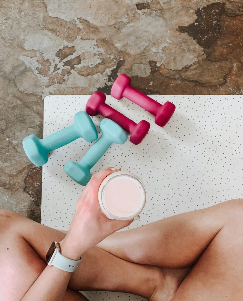 Pessoa sentada em um tapete de treino segurando um copo de whey protein, com halteres coloridos ao lado.