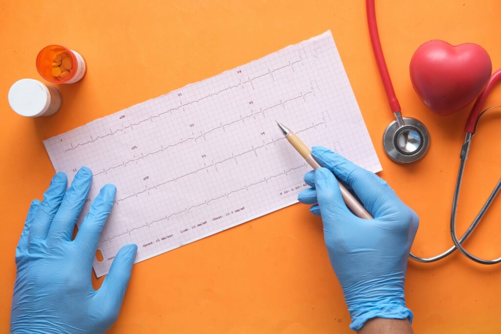 Profissional de saúde com luvas azuis analisando eletrocardiograma sobre fundo laranja, ao lado de remédios, estetoscópio e coração de borracha.