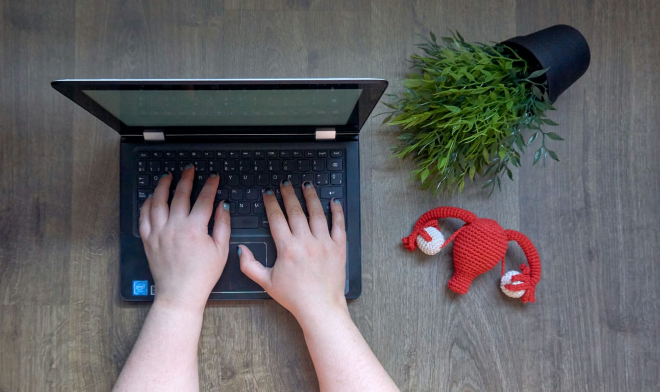 Mãos digitando em notebook com miniatura de útero de crochê ao lado, simbolizando pesquisa e informação sobre endometriose.