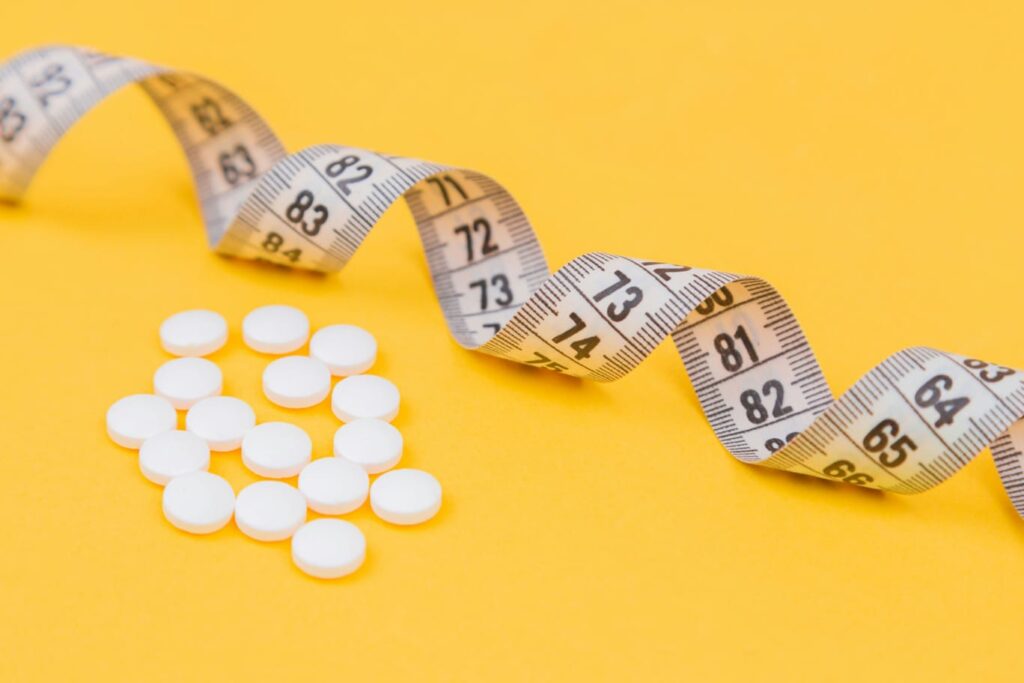 Comprimidos brancos ao lado de fita métrica em fundo amarelo, representando a perda de peso que medicamentos para emagrecer podem trazer.