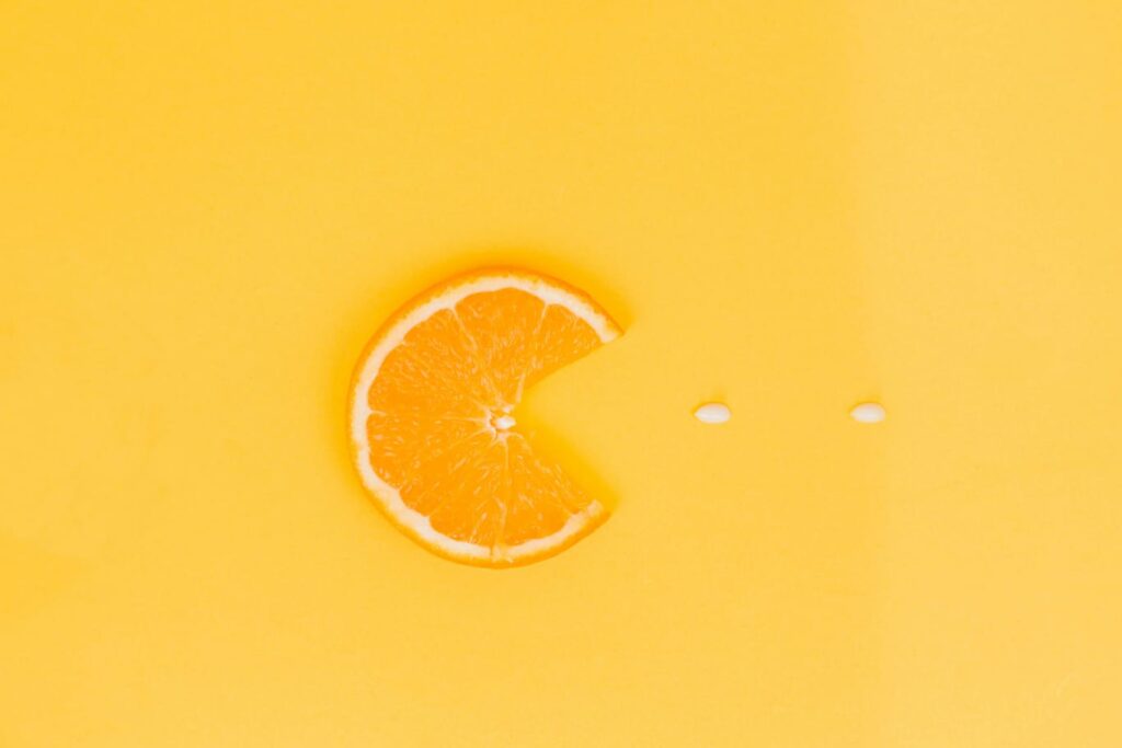 Fatia de laranja em formato de Pac-Man sobre fundo amarelo, com duas pílulas brancas de vitamina c à frente.