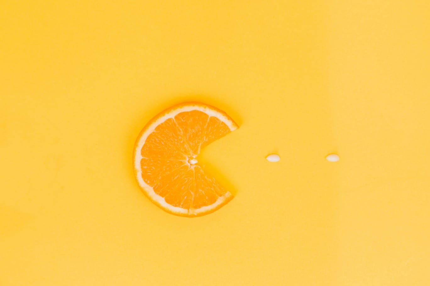 Fatia de laranja em formato de Pac-Man sobre fundo amarelo, com duas pílulas brancas de vitamina c à frente.