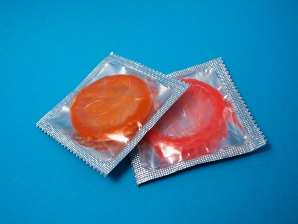 Dois preservativos coloridos em suas embalagens sobre um fundo azul, representando a importância do uso de camisinha na prevenção de infecções sexualmente transmissíveis.